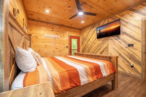 A wooden cabin room with a bed, a ceiling fan, a television, and a red door. - 🎱 Riversong Elegance | Game Room, Hot Tub & Sleeps 12, Minutes to Dollywood 🏔️ (Sevierville)