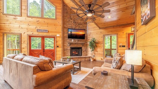 A log cabin interior with a fireplace, a ceiling fan, and a large window overlooking the forest. - 🎱 Riversong Elegance | Game Room, Hot Tub & Sleeps 12, Minutes to Dollywood 🏔️ (Sevierville)