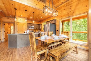 A wooden cabin interior with a dining area, kitchen, and large windows overlooking a forest. - 🎱 Riversong Elegance | Game Room, Hot Tub & Sleeps 12, Minutes to Dollywood 🏔️ (Sevierville)