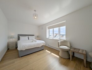 Ein Schlafzimmer mit Bett, Nachttischen, einem Sessel, einem kleinen Tisch, einem Fenster mit Blick und einer Deckenleuchte.