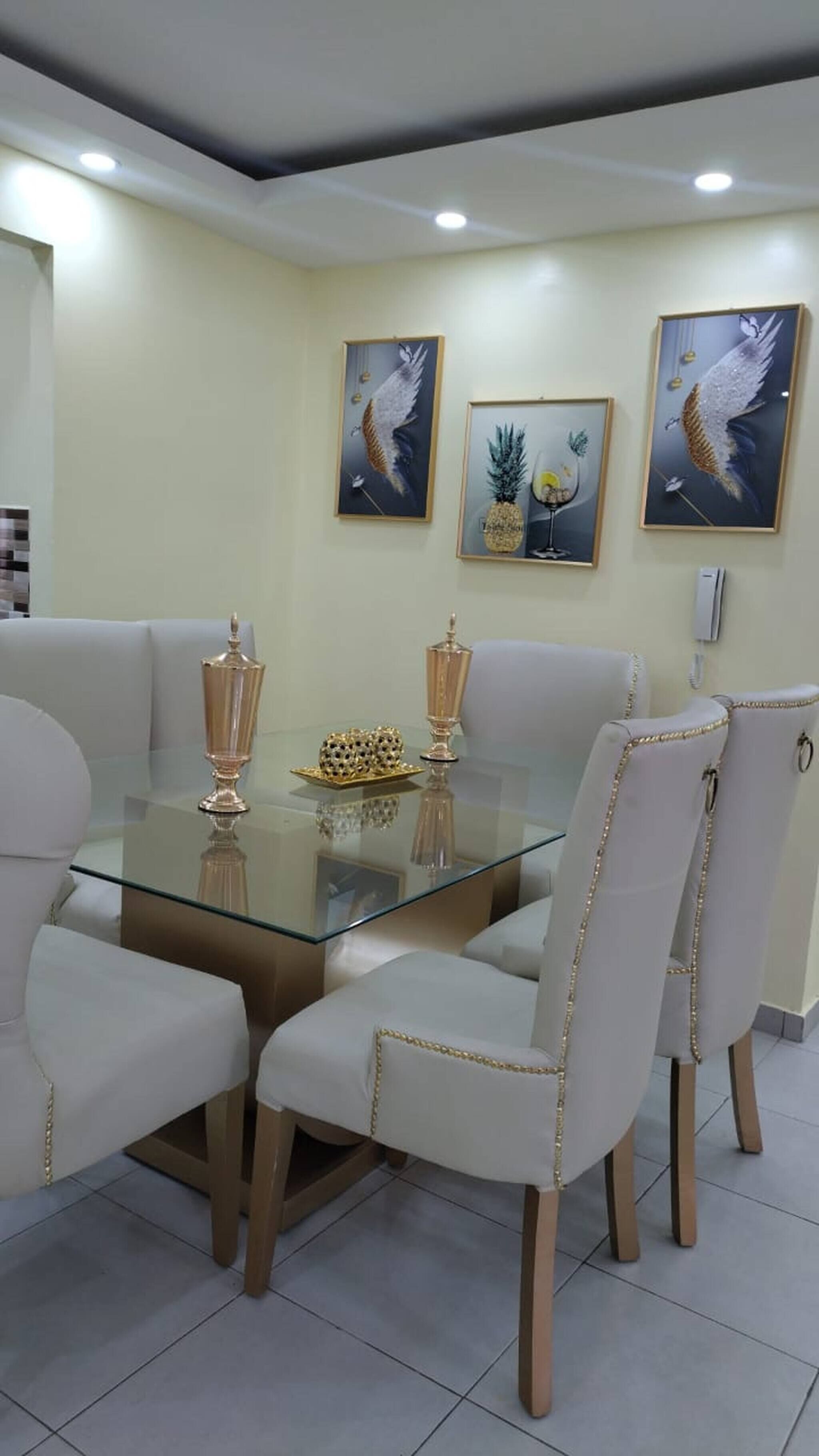 A dining area with a glass table surrounded by white upholstered chairs with gold trim. There are three framed artworks on the wall above the table.