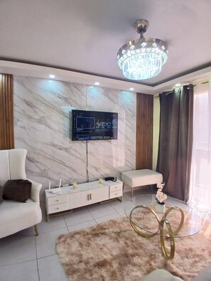 A modern living room with a marble wall, a mounted TV, a white sofa, a small side table, and a chandelier. - Beautiful 3 Bedroom Apartment With Pool on Av (Santo Domingo Norte)