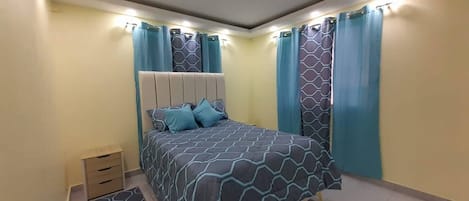 Un dormitorio con cama, cortinas, una mesita de noche y un pequeño mueble con cajones.
