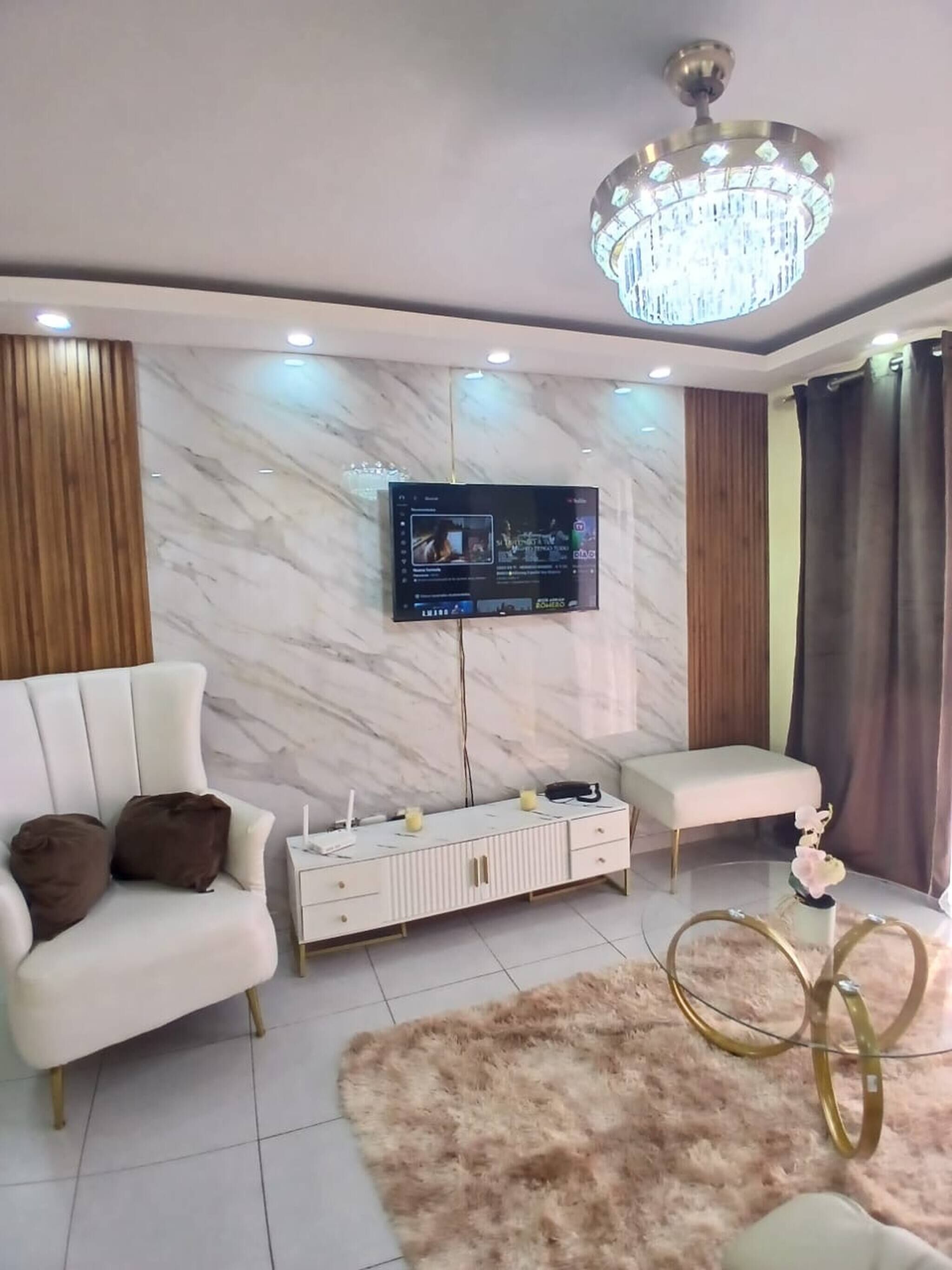Una sala de estar moderna con una televisión de pantalla plana montada en la pared, un sofá blanco, un sillón blanco, una alfombra mullida y un anillo decorativo dorado en el suelo.