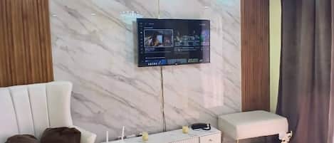 Una sala de estar moderna con una televisión de pantalla plana montada en la pared, un sofá blanco, un sillón blanco, una alfombra mullida y un anillo decorativo dorado en el suelo.