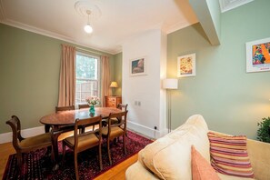 Ein Essbereich mit Holztisch und Stühlen, einem Fenster mit Vorhängen, einer Lampe und einer Couch mit Kissen.
