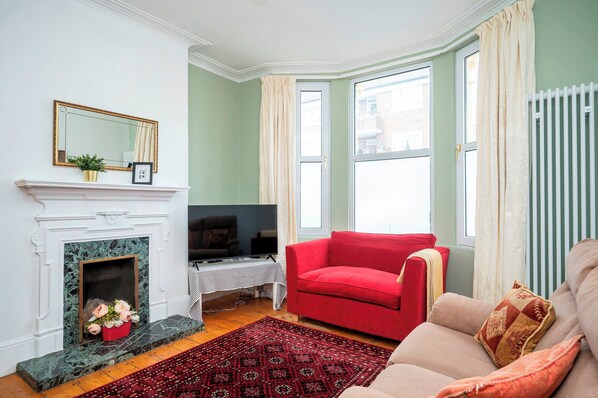 Una sala de estar con chimenea, un sofá rojo, un sofá beige, un televisor y un ventanal grande con cortinas.