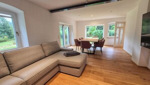 Een moderne woonkamer met een hoekbank, een eethoek met tafel en stoelen, en grote ramen met uitzicht op groen.
