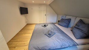 Een slaapkamer met een bed, kussens, een flatscreen televisie aan de muur en een houten vloer.