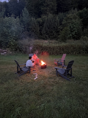 A group of people sitting on Adirondack chairs around a campfire in a grassy outdoor area with trees in the background.