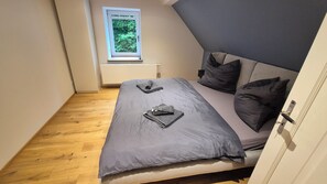 Una cama pequeña y bien hecha, con ropa de cama y almohadas grises, suelo de madera, una ventana con vistas a la vegetación y una puerta blanca.