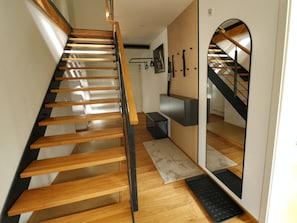 A wooden staircase with a black handrail, leading to a landing with a black mat and a wall-mounted shelf.