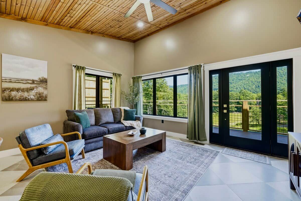 A spacious living room with a sofa, armchairs, a coffee table, and large windows offering a view of greenery.