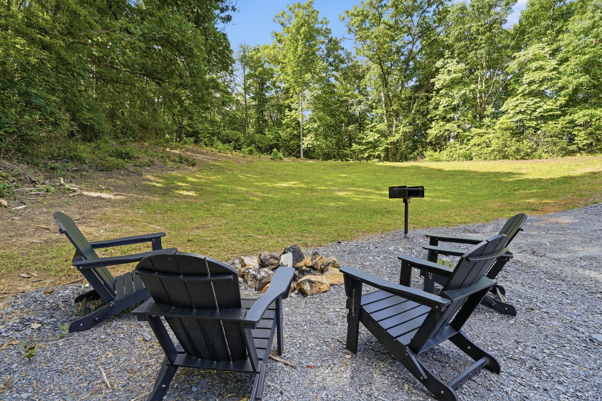 Two Adirondack chairs facing a fire pit with a signpost in a gravel area surrounded by trees.