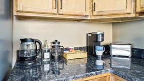 A kitchen with wooden cabinets, a coffee maker, a kettle, a coffee pot, a glass carafe, a bottle of French vanilla, a coffee grinder, a basket of snacks, and a toaster. - Lovely 1-bedroom cabin with a hot tub in the mountains of Elizabethton (Elizabethton)