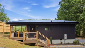 A single-story building with a dark exterior, a covered porch with a wooden staircase, and a small garden area. - Lovely 1-bedroom cabin with a hot tub in the mountains of Elizabethton (Elizabethton)