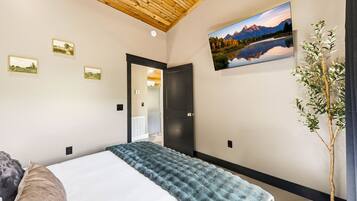 A bedroom with a bed, a wooden ceiling, a painting of a landscape, and a potted plant.