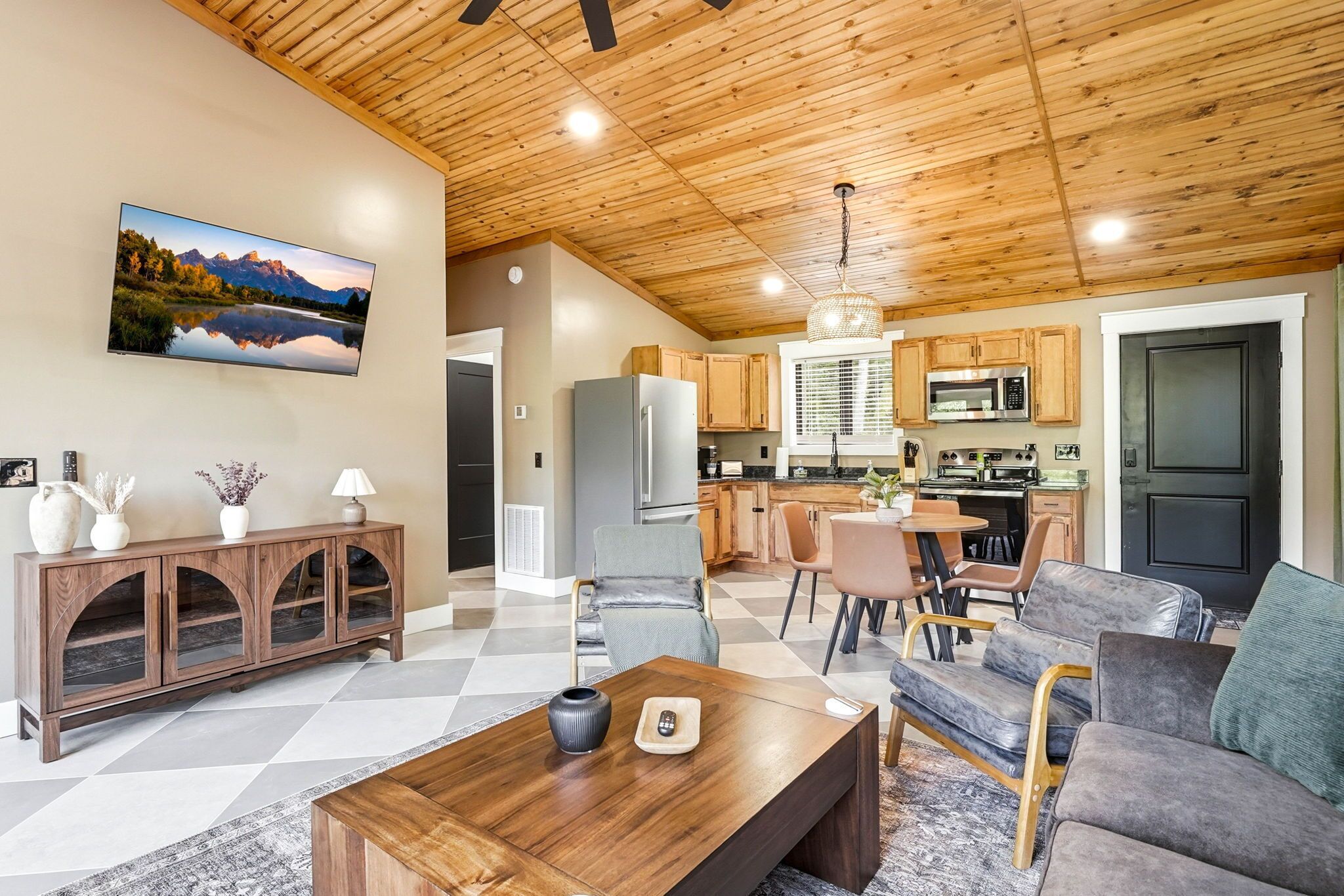 A modern living room with a wooden ceiling, a large flat-screen TV, a dining area with a table and chairs, and a kitchen with wooden cabinets and appliances.