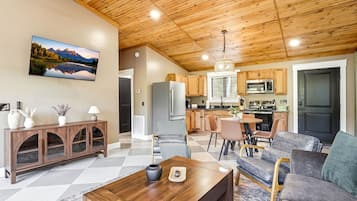 A modern living room with a wooden ceiling, a large flat-screen TV, a dining area with a table and chairs, and a kitchen with wooden cabinets and appliances.