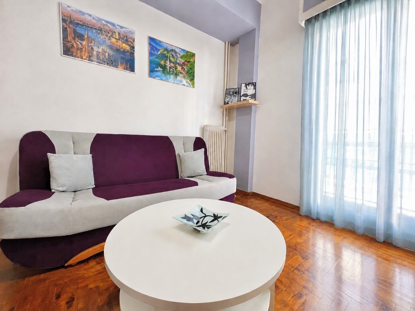 A living room with a purple and white sofa, a white coffee table, and two paintings on the wall.