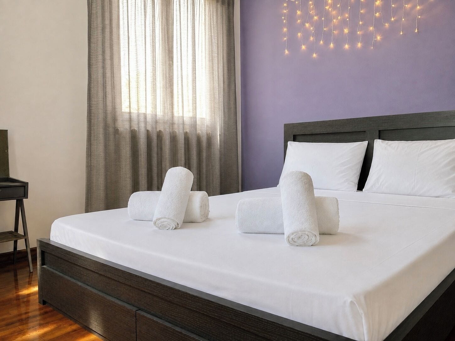 Een net opgemaakt bed met witte lakens en opgerolde handdoeken, een houten hoofdbord en een muur met lichtslingers.