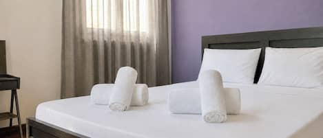 Una cama bien hecha con sábanas blancas y toallas enrolladas, cabecera de madera, y una pared con luces colgantes.