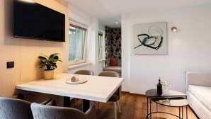 A modern interior with a dining area, a flat-screen TV, a painting, and a small table with a bottle and glasses. - Grappeskopf 23 Apartment (Gaschurn)