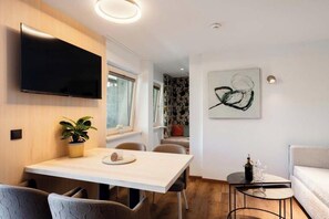 A modern interior with a dining area, a flat-screen TV, a painting, and a small table with a bottle and glasses. - Grappeskopf 23 Apartment (Gaschurn)