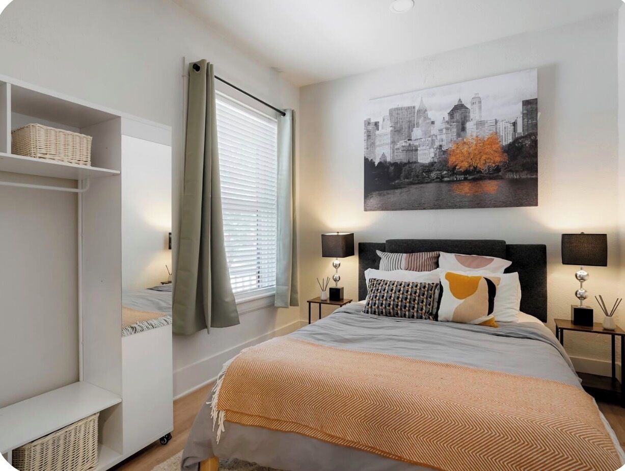 A bedroom with a bed, a cityscape painting, a nightstand, a lamp, and a window with curtains.
