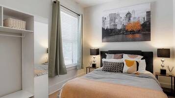 A bedroom with a bed, a cityscape painting, a nightstand, a lamp, and a window with curtains.