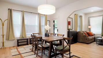 A modern living room with a dining area, featuring a wooden table, chairs, a sofa, and a large window with curtains.