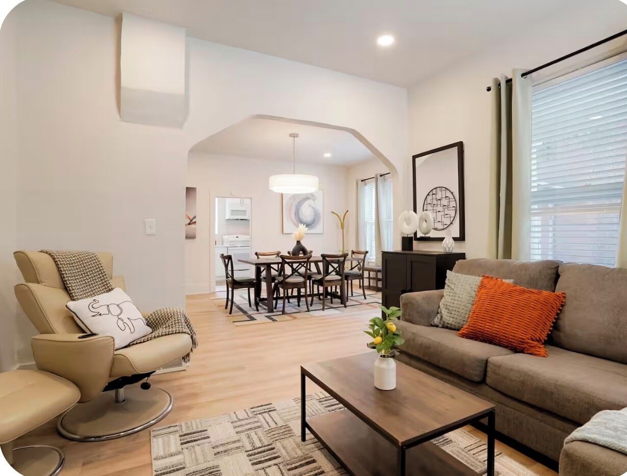A modern living room with a sofa, armchair, coffee table, and a dining area with chairs and a table.