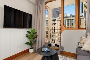Ein modernes Wohnzimmer mit einem Flachbildfernseher, einer Topfpflanze, einem runden Tisch mit einem dekorativen Gegenstand und Blick auf die Skyline durch ein Fenster.