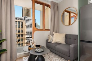 Ein modernes Wohnzimmer mit einem grauen Sofa, einem runden Spiegel und Blick auf eine Stadtlandschaft durch große Fenster.