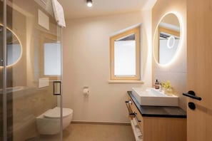A modern bathroom with a glass shower enclosure, a toilet, a sink with a mirror, and a window. - Apartment Piz Buin 31 (Gaschurn)