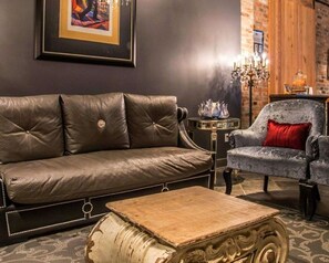 A leather sofa, a patterned armchair, a small table, and a decorative coffee table in a room with a brick wall and a chandelier.