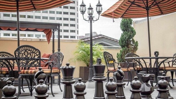An outdoor seating area with chess pieces arranged on a checkered floor, surrounded by tables and chairs with striped umbrellas.