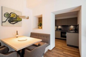 Eine moderne Küche mit dunklen Schränken, ein Essbereich mit einem Holztisch und grau gepolsterten Stühlen sowie ein an der Wand hängendes abstraktes Gemälde.