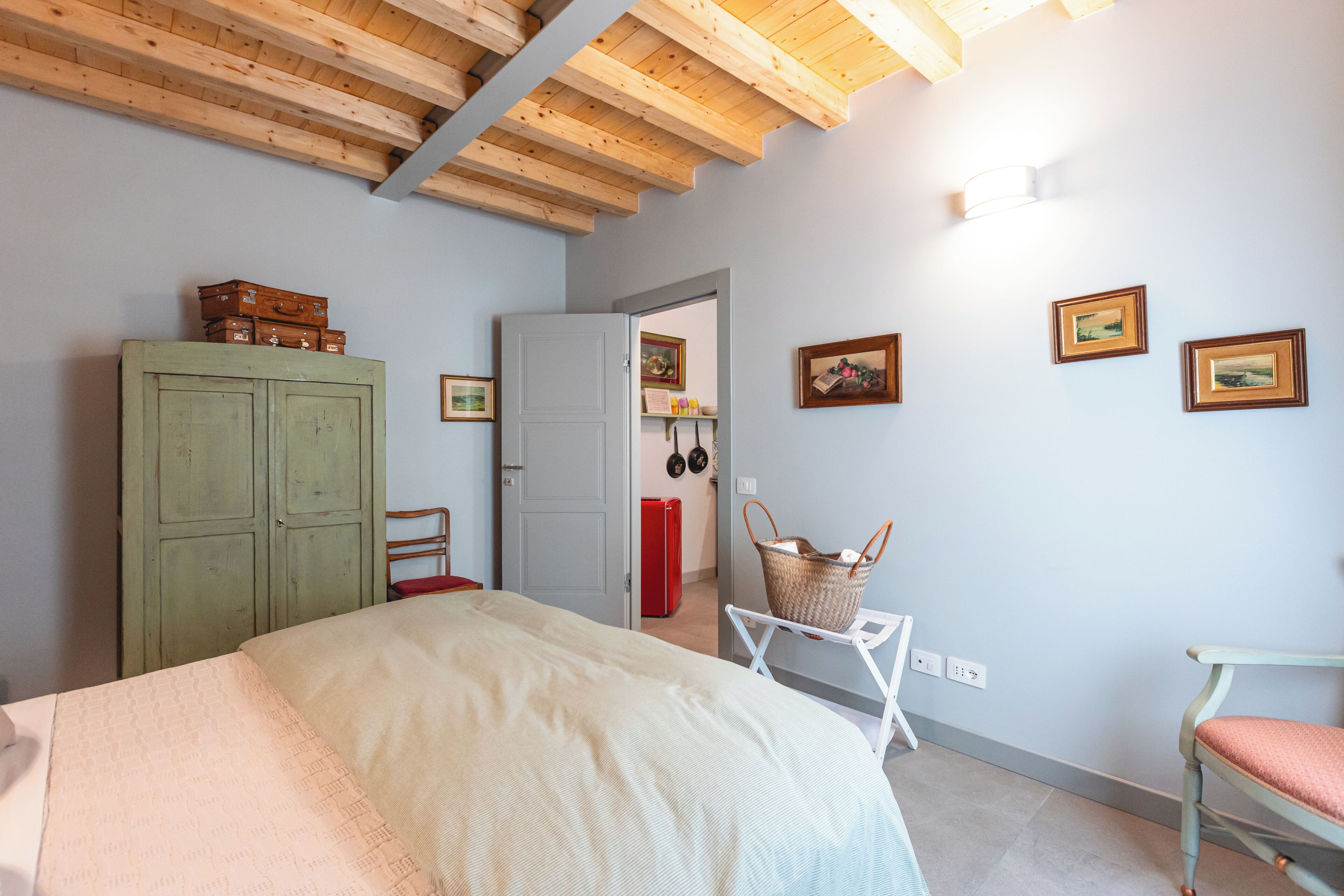 A bedroom with a bed, a wardrobe, a chair, a small table with a basket, and a door leading to another room.