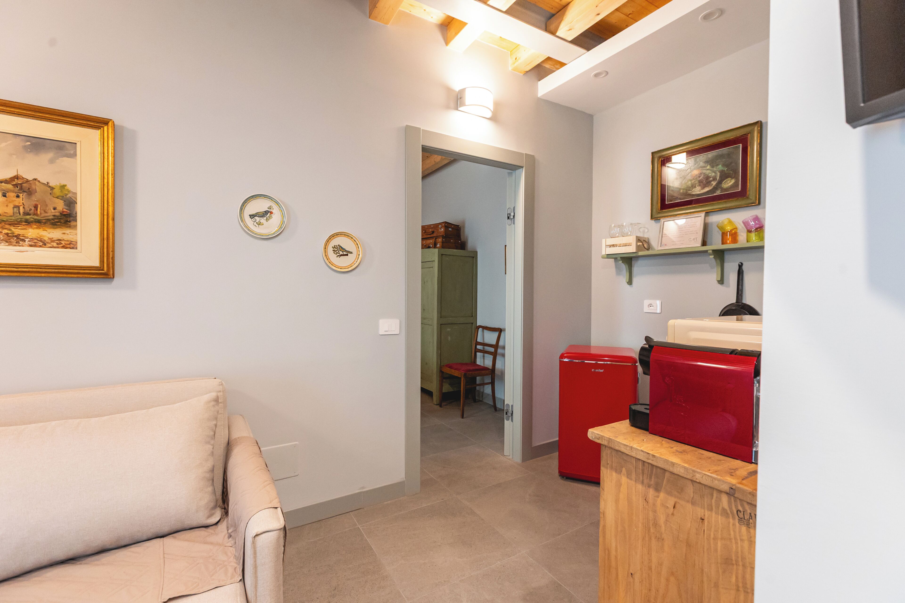 Une pièce avec un canapé beige, un réfrigérateur rouge et un meuble en bois. Il y a deux luminaires fixés au mur et un tableau encadré accroché au mur.