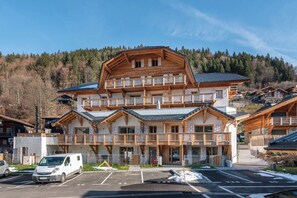 Un immeuble en bois à plusieurs étages, doté de balcons, d’un parking et situé dans un paysage vallonné et boisé.