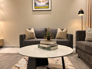 A modern living room with a grey sofa, a round coffee table, and a framed landscape painting above. - Rubis Signature Suites (Abuja)
