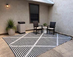 Un espacio al aire libre con dos sillas, una mesa pequeña, una planta en maceta y un contenedor de basura.