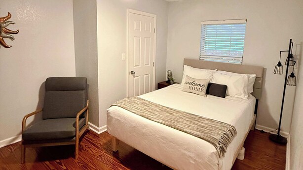 A bedroom with a bed, a chair, a door, a window, and a floor lamp. - 4BR Near Alamodome & Pearl | Sleeps 10 (San Antonio)