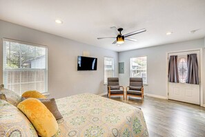 Un dormitorio con cama, dos sillones, ventilador de techo, televisor y ventanas amplias.