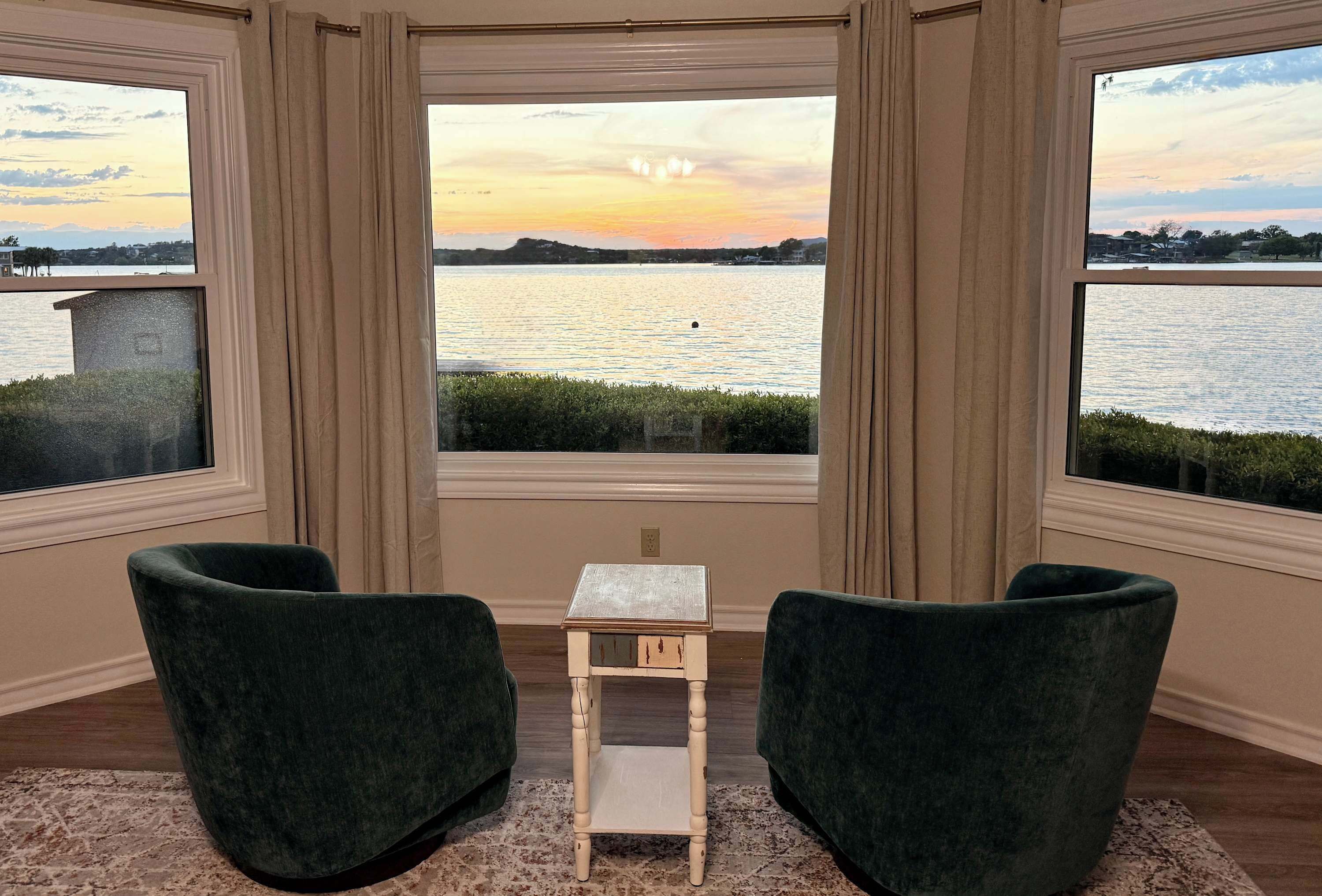 Et værelse med udsigt over en sø i solnedgang, to grønne stole og et lille træbord.