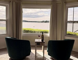 Ein Zimmer mit Blick auf ein Gewässer, zwei Stühle, ein kleiner Tisch mit einer Weinflasche und einem Glas sowie ein Fenster mit Vorhängen.