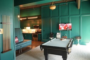 A billiards table in a room with teal walls, a kitchen area, and a television displaying a sports channel.