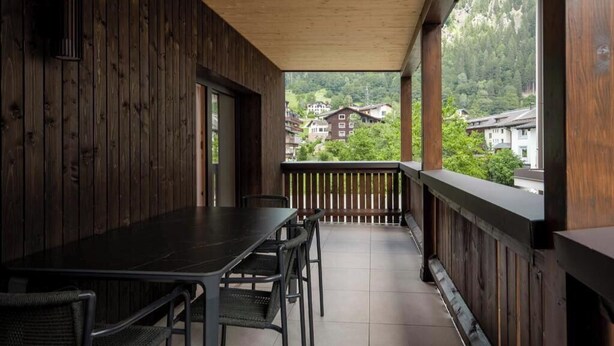 A wooden balcony with a table and chairs, overlooking a mountainous area with buildings. - Tafamunt 11 Apartment (Gaschurn)