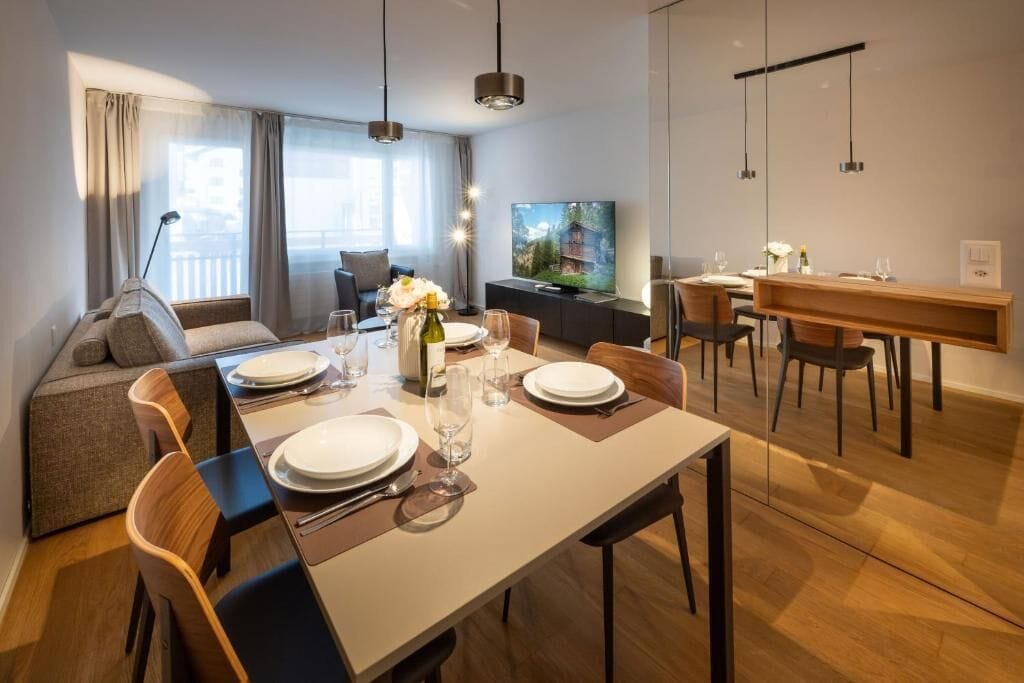 Un espace de repas moderne avec une table dressée pour quatre personnes, un canapé et un téléviseur en arrière-plan.